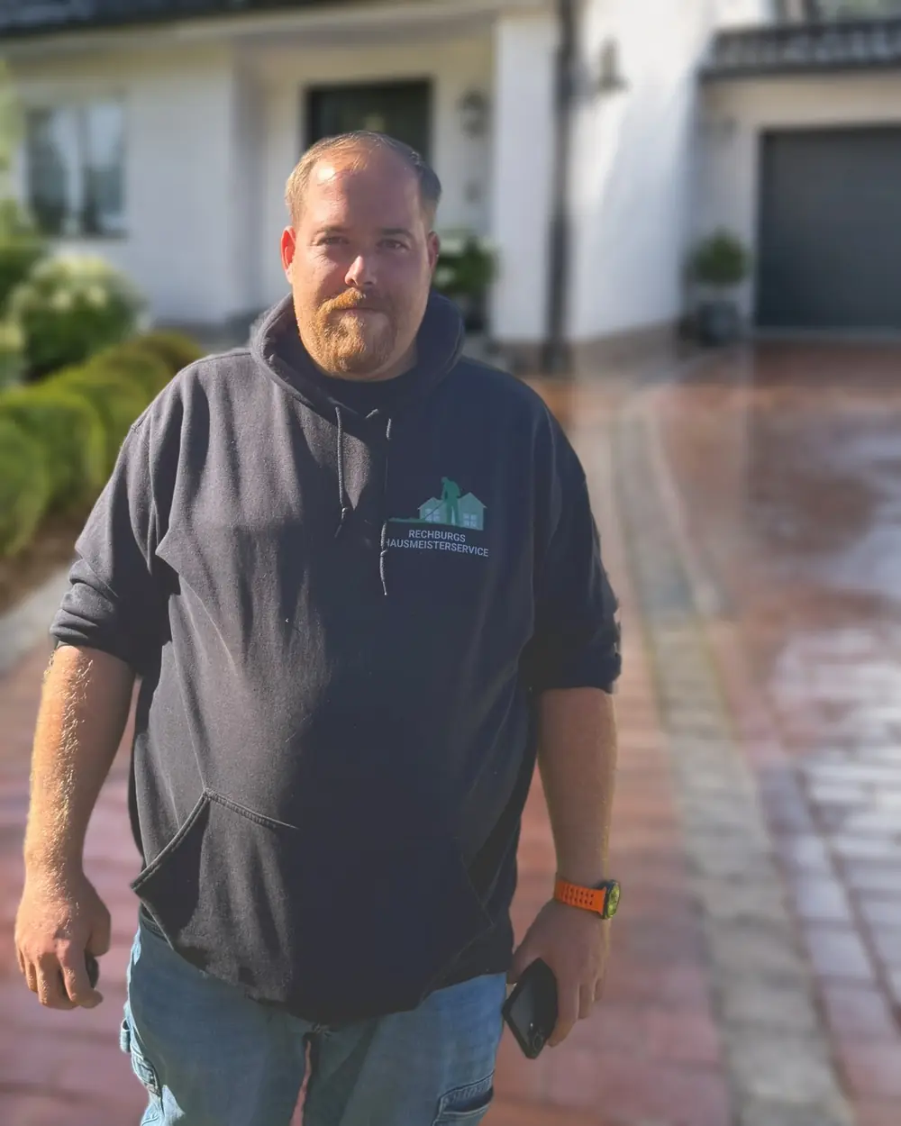 Pascal Rechburg, Inhaber von Rechburgs Hausmeisterservice, im Firmen-Hoodie; der Hintergrund mit Wohnhaus und Einfahrt ist KI-generiert